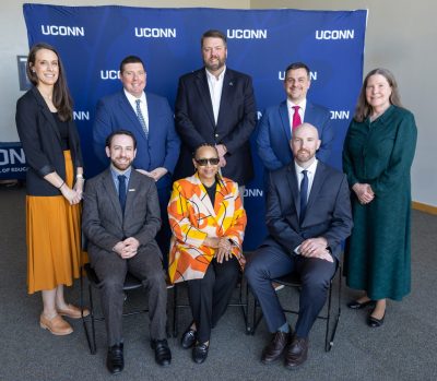 Three women and five men post in a group in front of a blue backdrop that repeats "UConn" in white.
