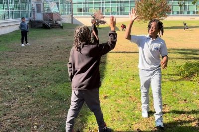 Two middle-school-age Black males high five outside of a school in a field.