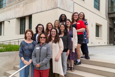 Eleven women stand on the exterior steps of the building in two rows and smile at the camera.