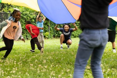 A Black college student squats down while holding an edge of a large parachute over their head outside in a field. Elementary school children also hold the parachute or run underneath it.