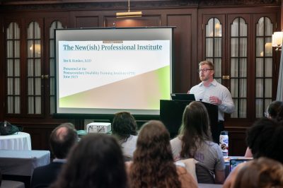 A white man stands at a podium at the front of a room full of seated audience members, while a projector screen behind him displays slides.