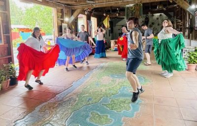 College students, some wearing brightly colored skirts, dance inside a room.